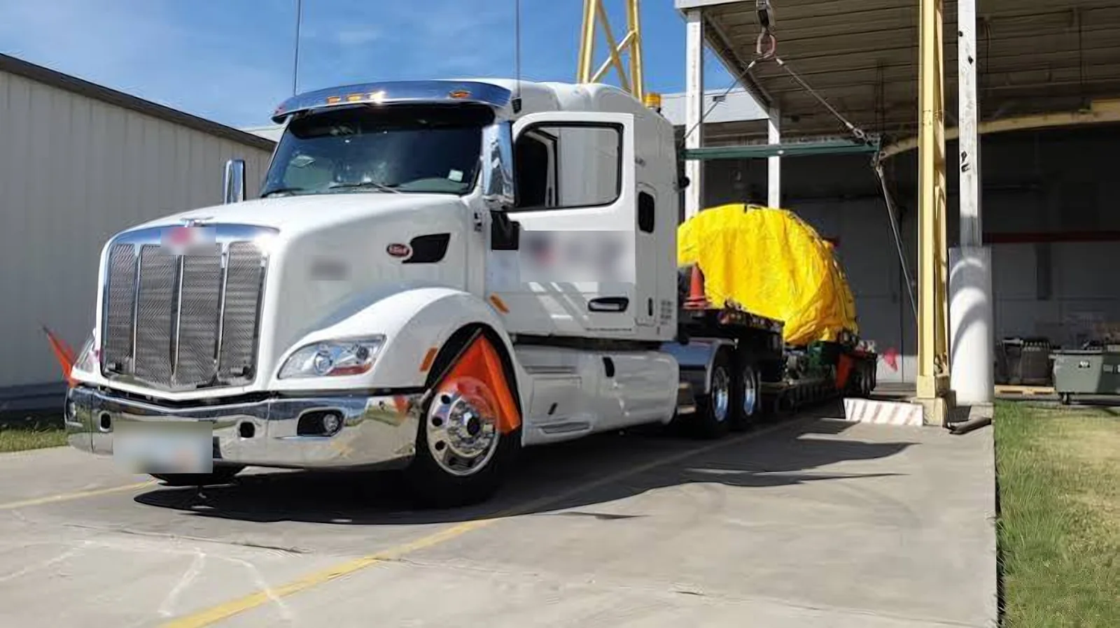 White semi truck pulling a step deck with yellow tarped aerospace cargo out of a hangar loading bay