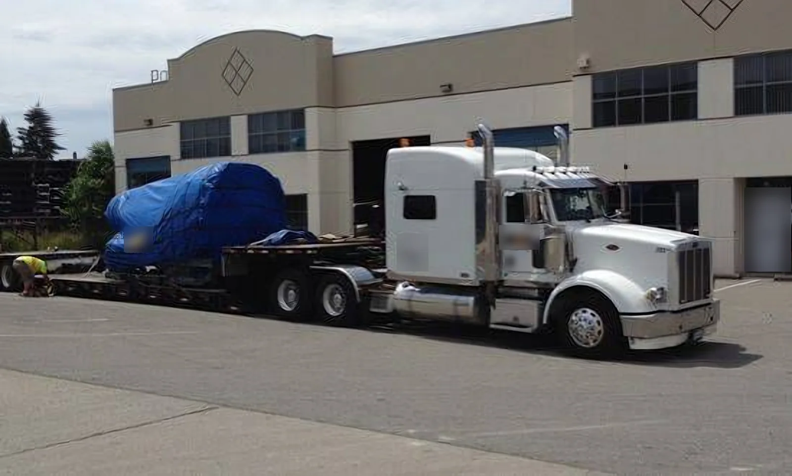 White semi truck pulling a double drop trailer with a blue tarped aerospace load outside an industrial building