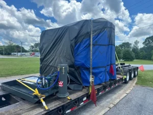 Machine covered in black and blue tarps on a step deck