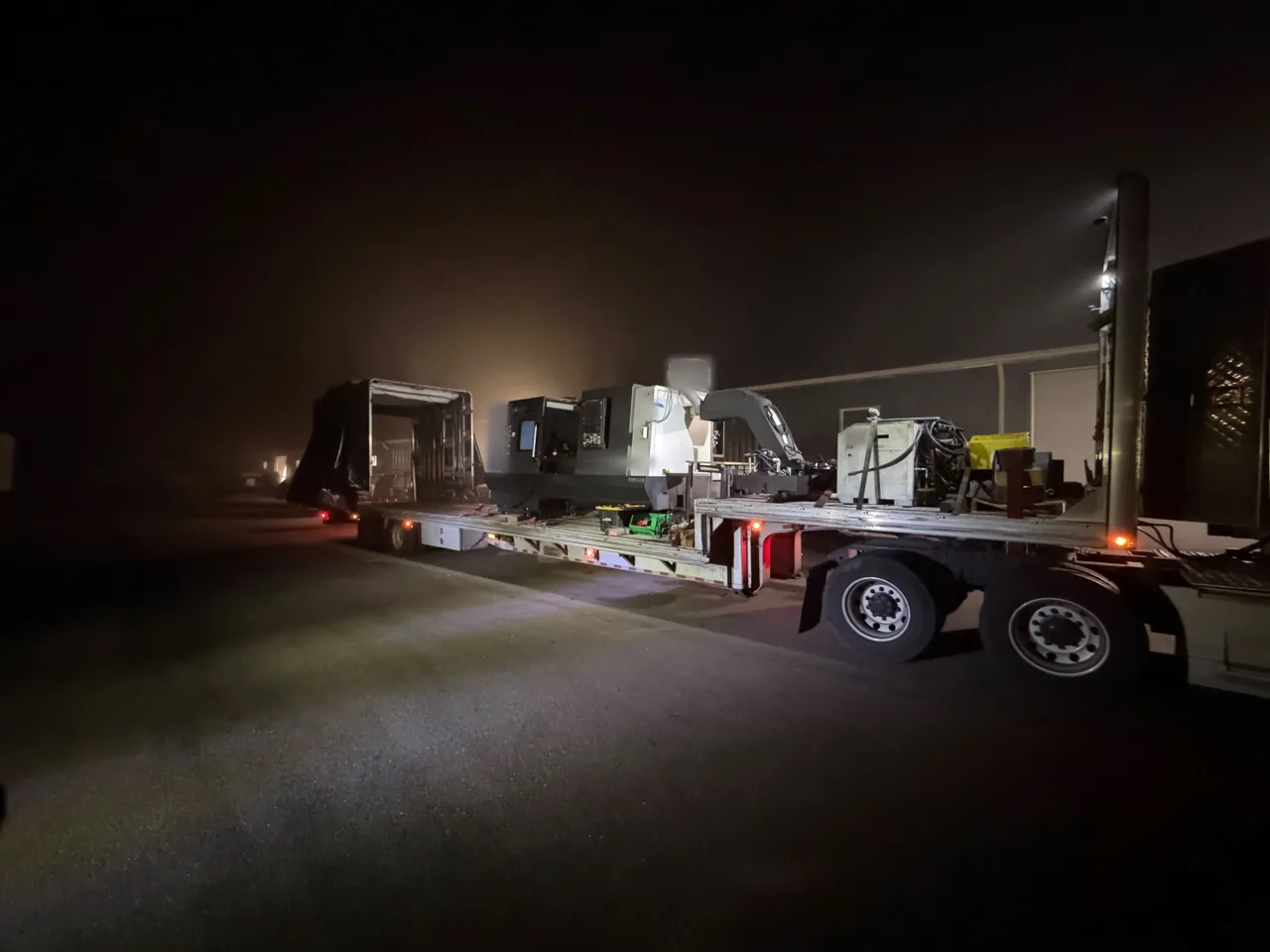 CNC lathe and chip conveyor loaded on the lower deck of a step deck Conestoga at night, tarp kit open at the rear