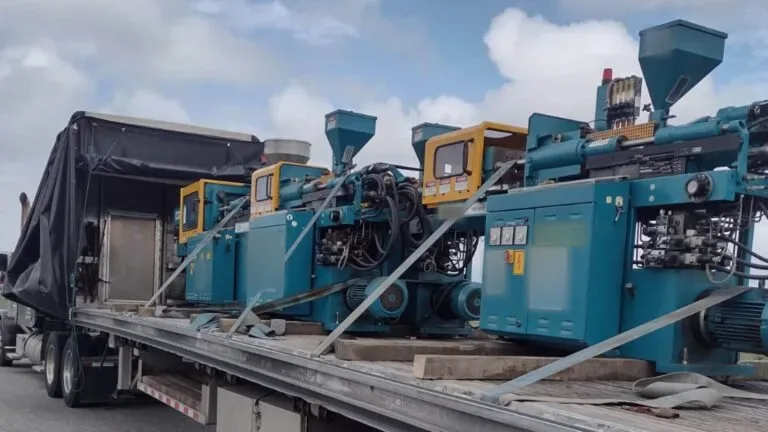 Conestoga machinery transport Los Angeles: four teal injection molding machines on a flatbed Conestoga, tarp rolled forward