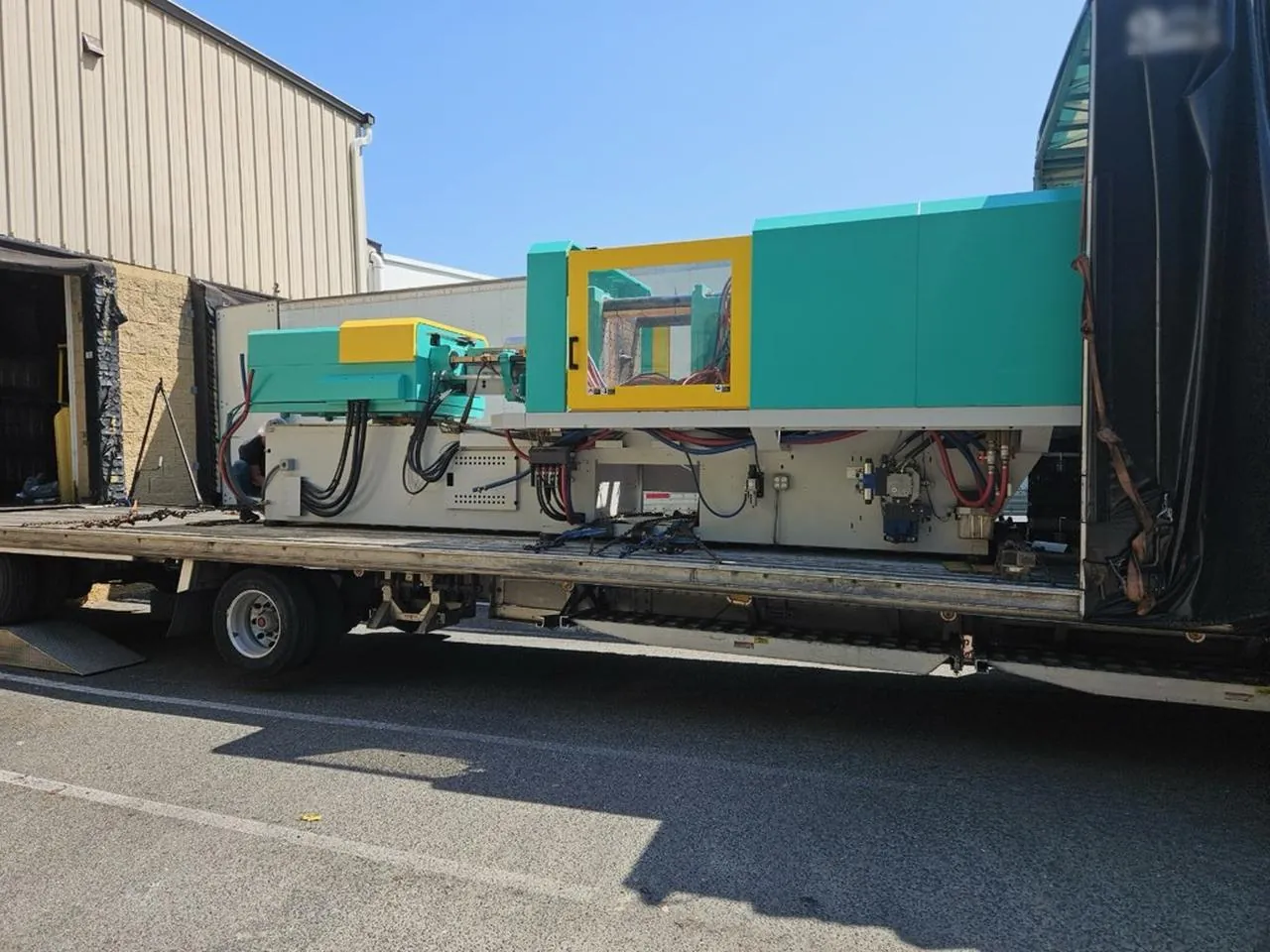 Injection molding machine on a Conestoga trailer deck beside a warehouse dock with the tarp rolled back