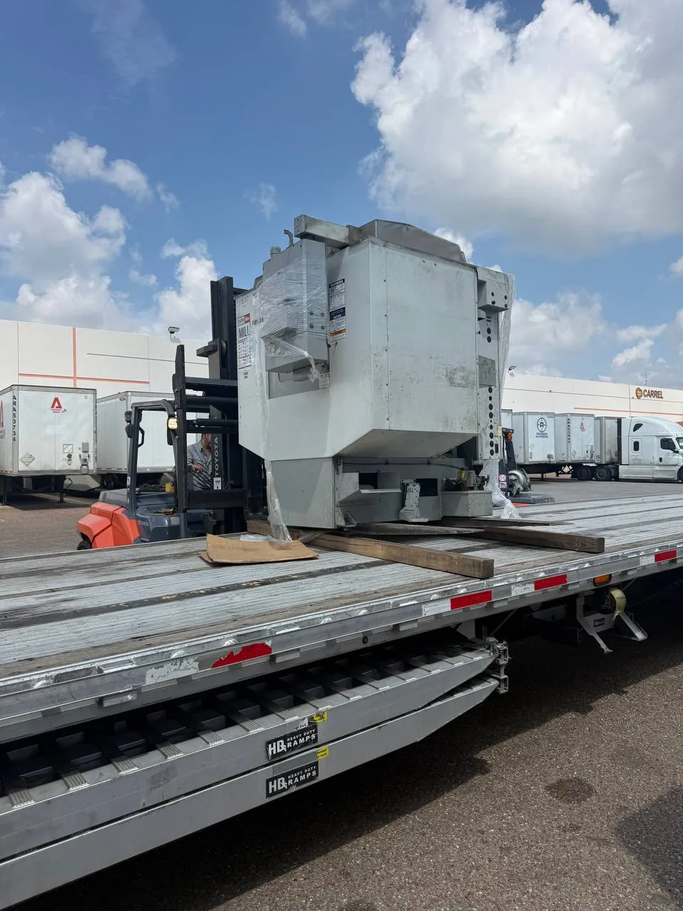 Forklift raising a Haas Mini Mill off wood dunnage on a step deck trailer, loading ramps visible below