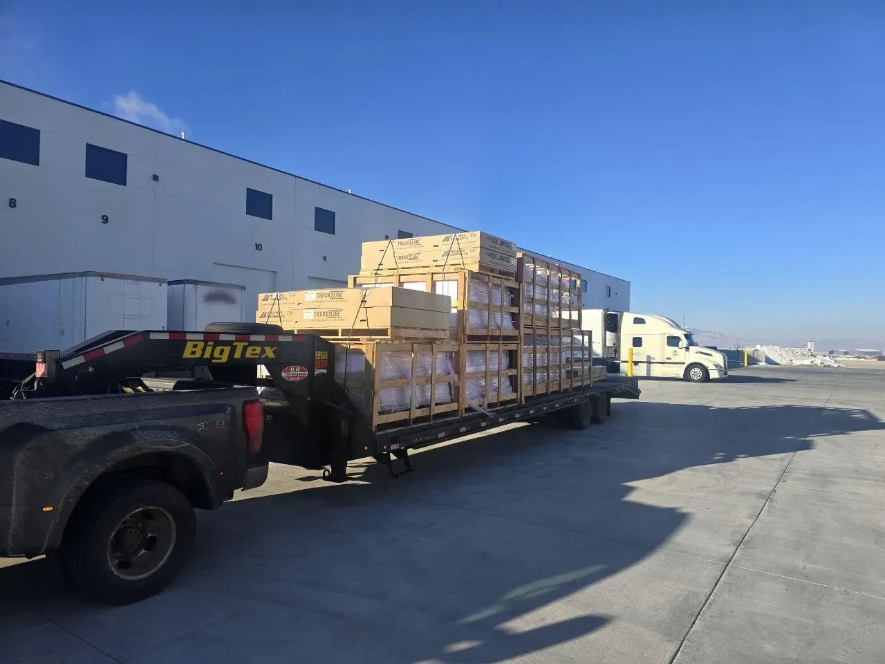 Dually pickup truck pulling a gooseneck flatbed trailer loaded with stacked wooden crates at a warehouse