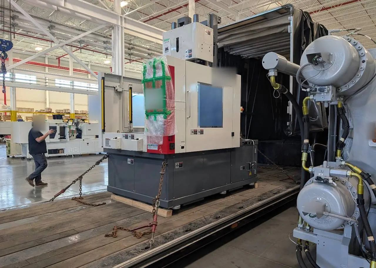 Injection molding machine on a wood skid, chained at its base inside an open Conestoga at a plant dock