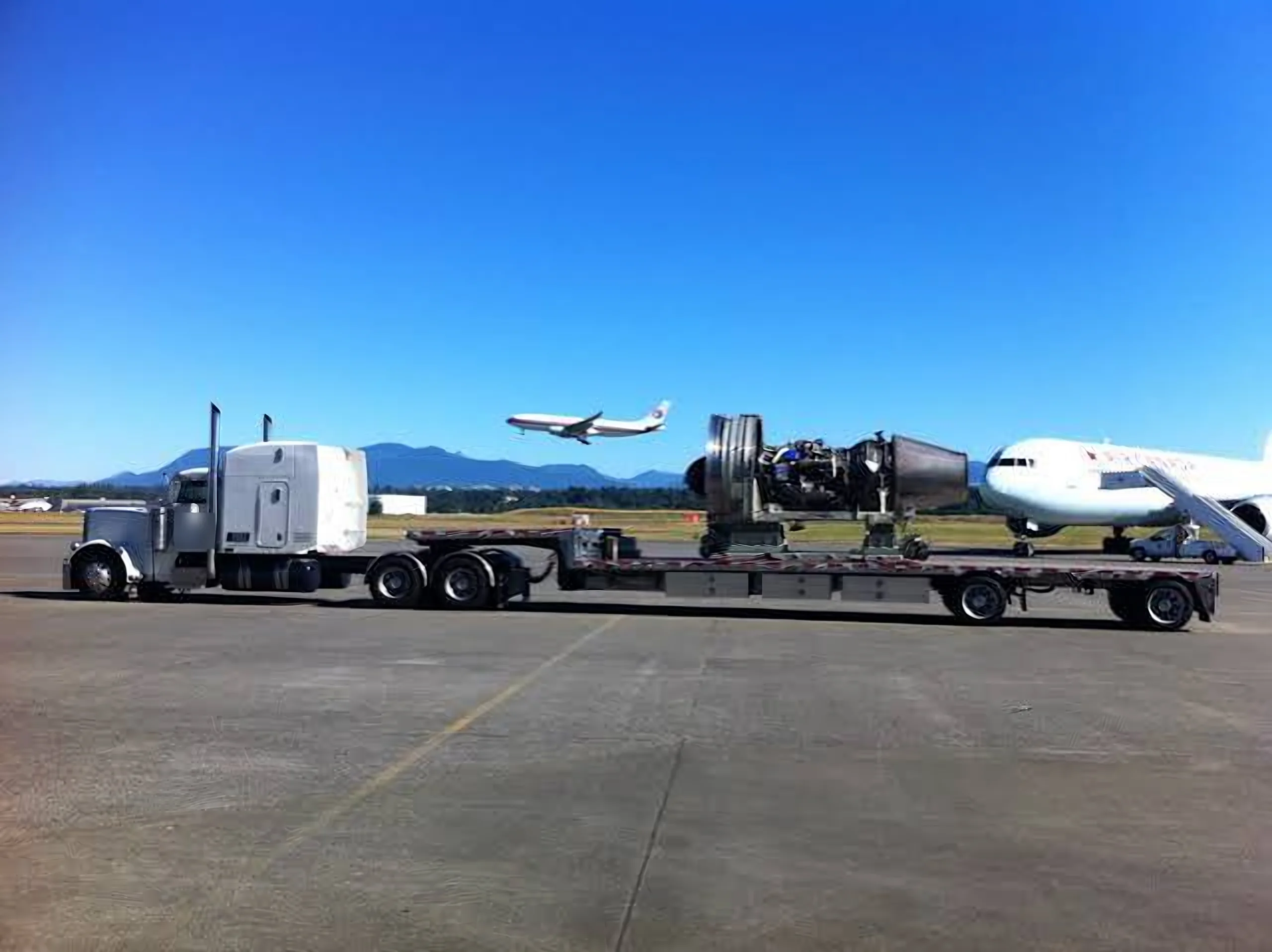 Jet engine on a stand chained to a step deck trailer behind a white semi truck on an airport ramp, with airliners nearby