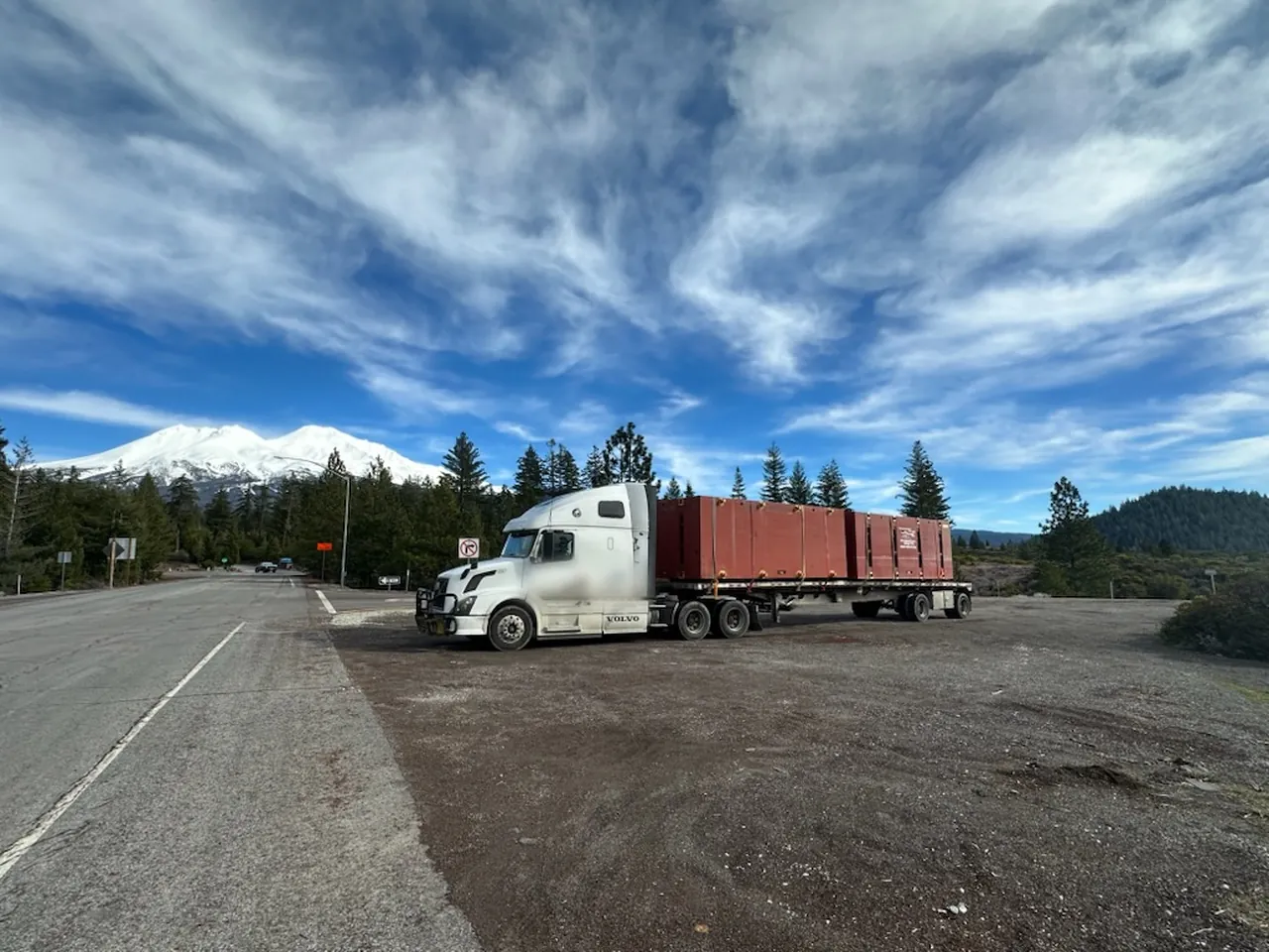 White semi with a loaded flatbed parked at a mountain roadside pullout under a wide blue sky