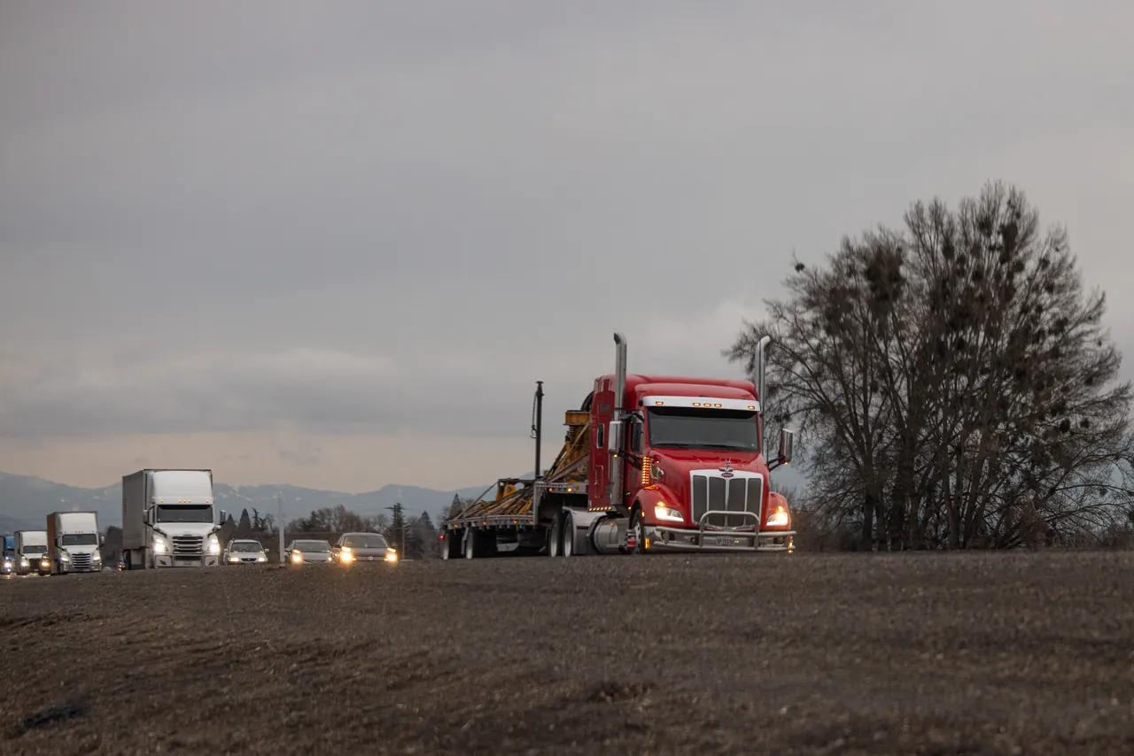 Red tractor pulling a loaded step deck trailer on a highway with trucks and cars behind