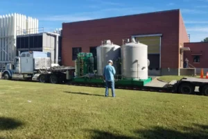 Natural gas compressor shipping: green skid with two grey vessels on a step deck lowboy beside a brick plant