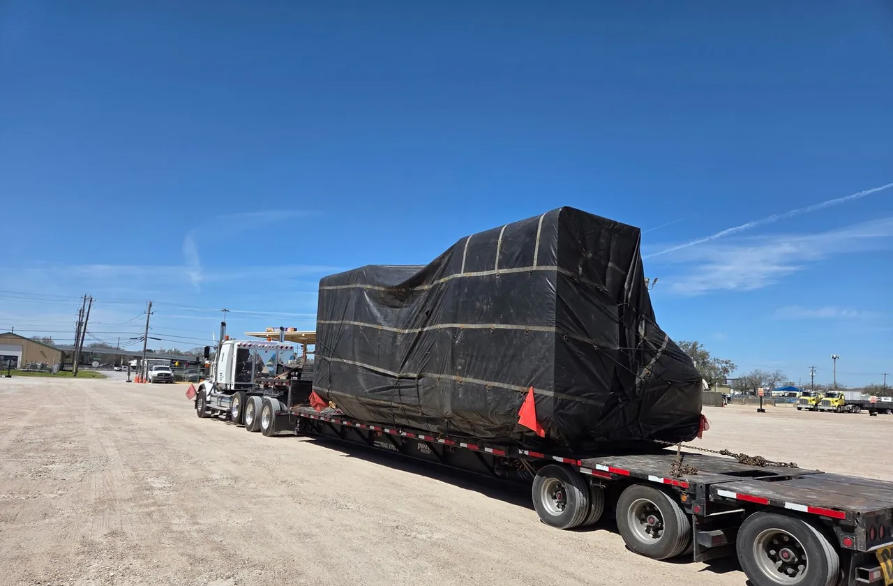 Large tarped machine chained to a step deck trailer with red corner flags, staged in a gravel yard