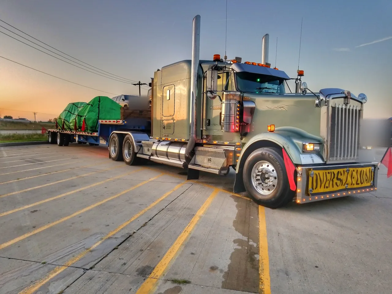 Semi truck with an OVERSIZE LOAD banner and red flags pulling a step deck with green tarped freight