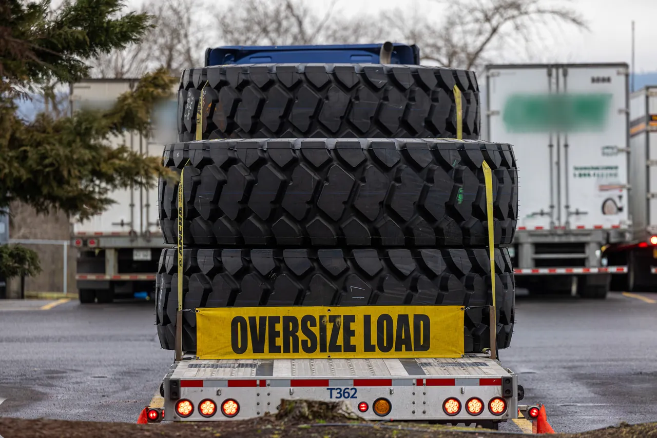 Stack of large off-road tires strapped to a step deck trailer with an oversize load banner on the back