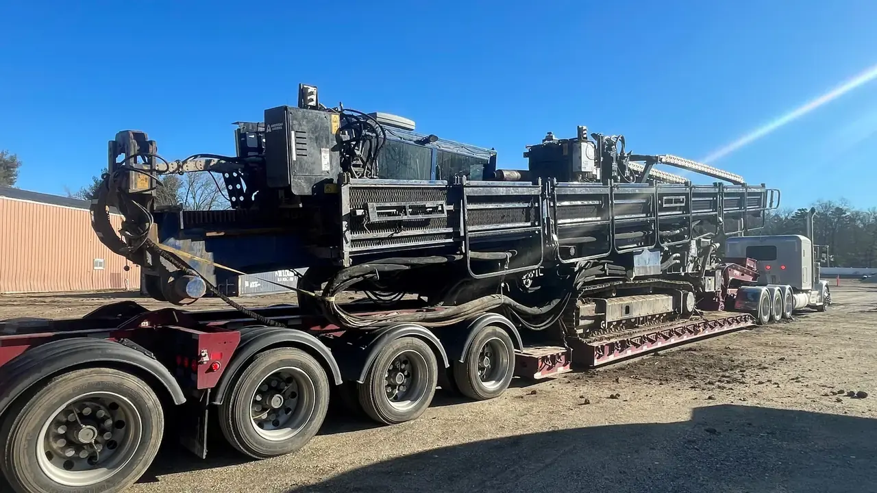 Heavy tracked drilling equipment loaded on a multi-axle lowboy trailer behind a gray semi