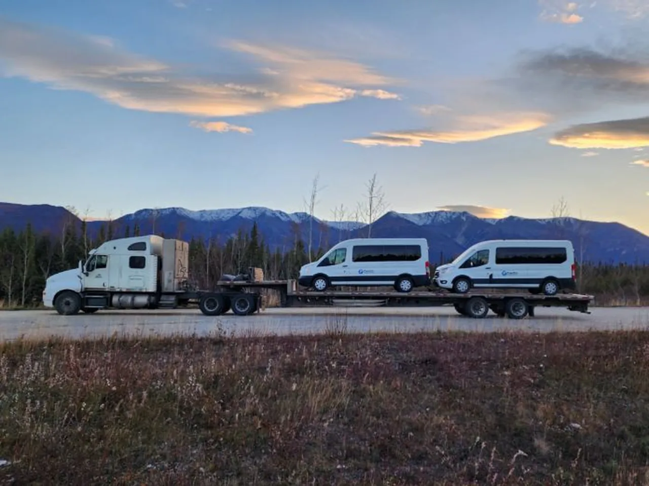 Two white cargo vans loaded on a step deck trailer behind a white semi at sunset