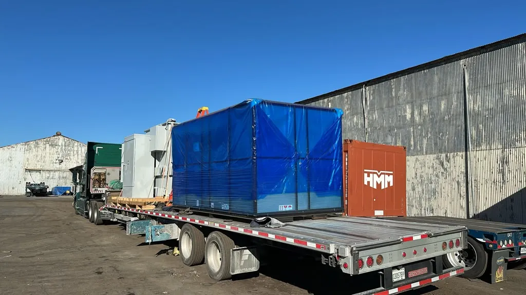 Shrink wrapped machine crate secured on a step deck trailer parked at a Los Angeles area warehouse