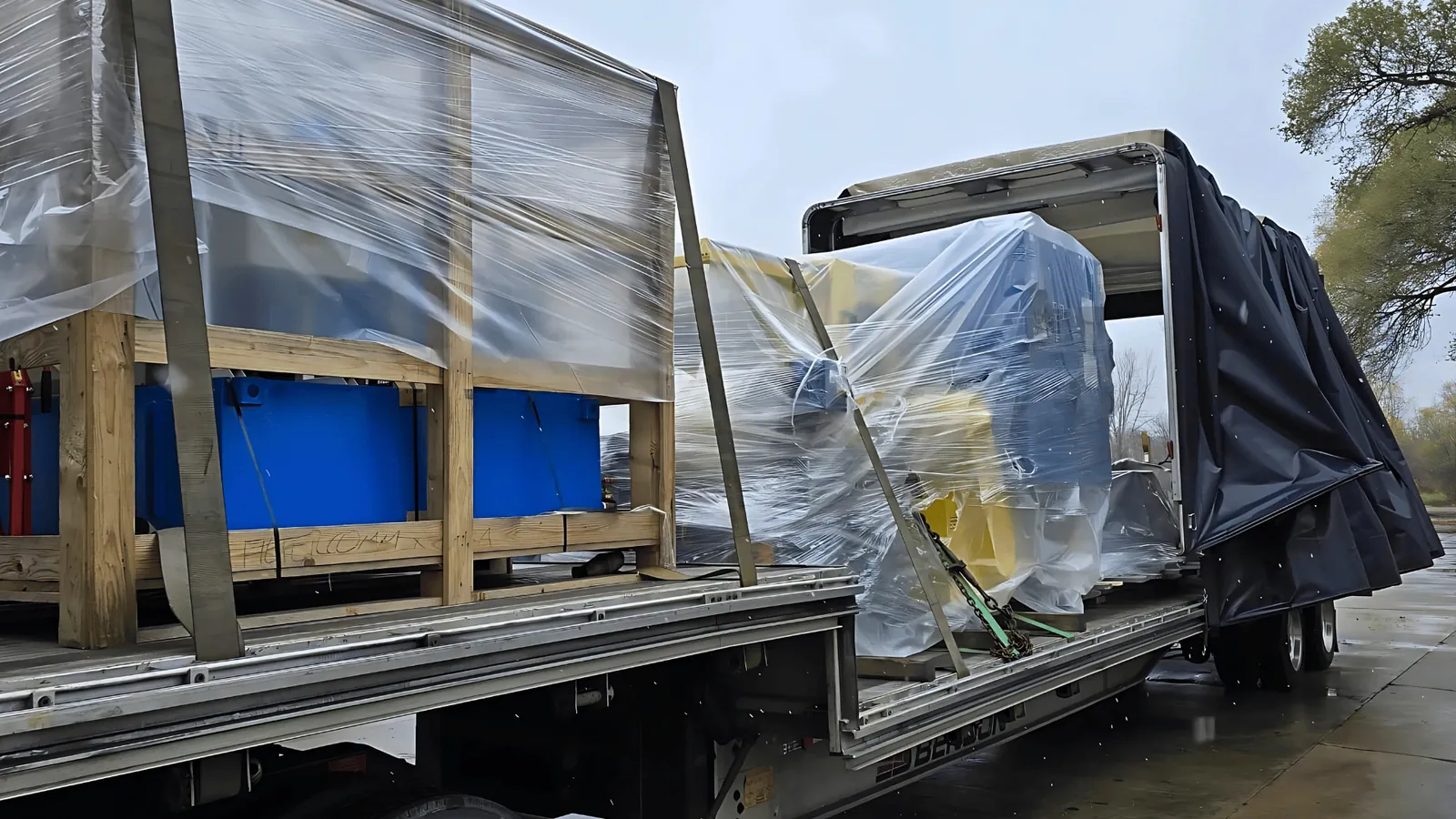 Shrink wrapped machinery and crates under the open cover of a Conestoga trailer on a rainy day