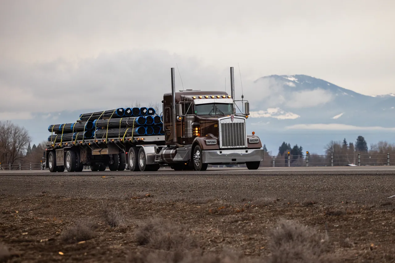 Semi with a flatbed loaded with bundled steel pipe parked near a highway with a snowy mountain behind