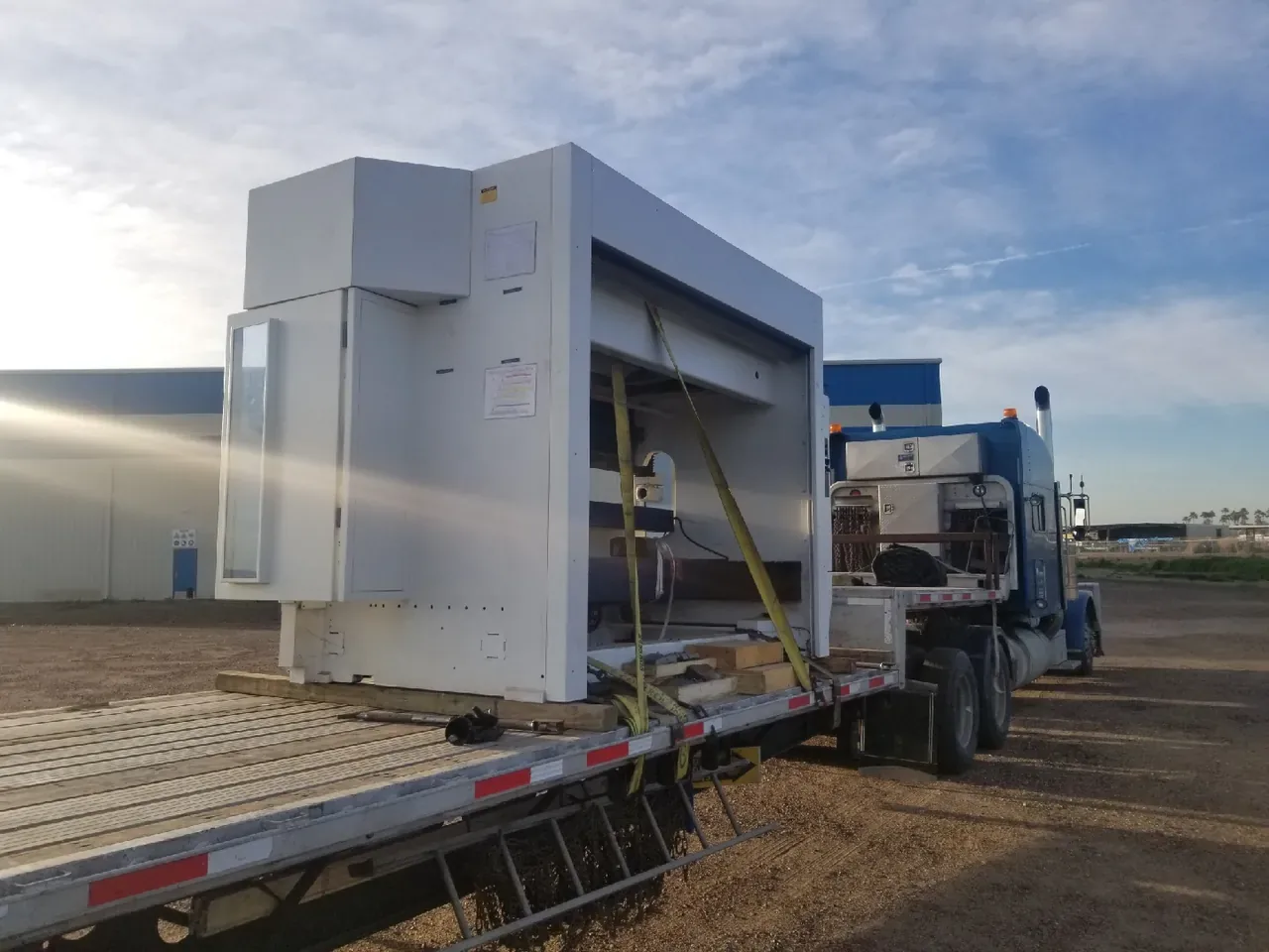 Tall white equipment enclosure strapped on the lower deck of a step deck trailer behind a blue tractor