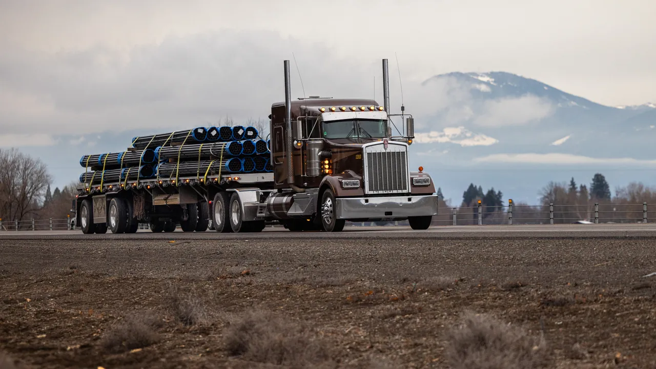 Flatbed truck hauling a load of blue pipe on a highway with mountains behind