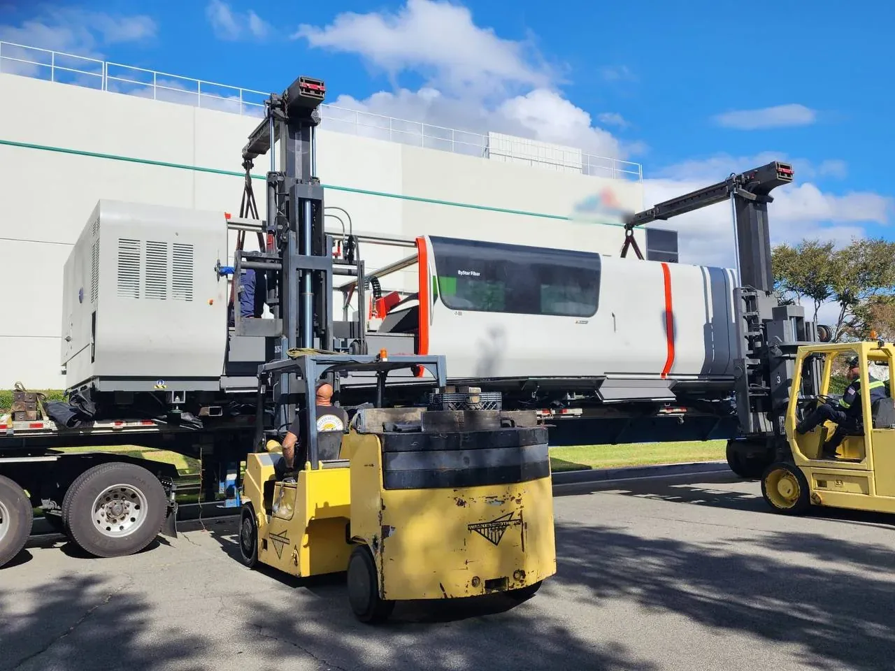 Two forklifts unloading a heavy machine from a flatbed at delivery