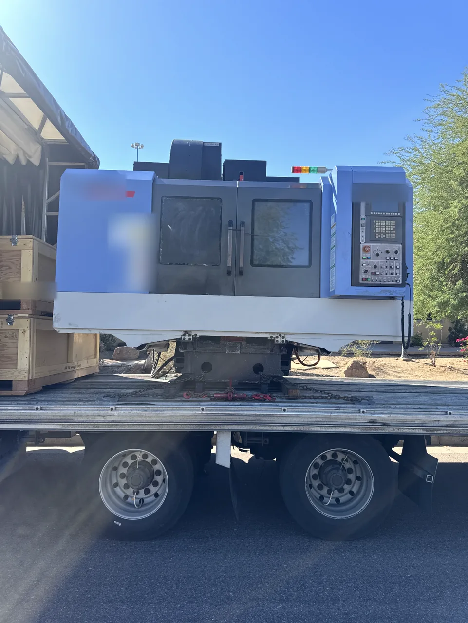 Vertical machining center chained at its base on a Conestoga deck, with the tarp rolled back and crates beside it